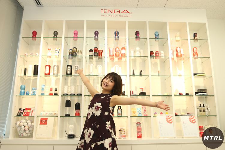 株式会社TENGAのオフィスにあるプレスルームで歴代の人気商品が並ぶディスプレイの前でポーズをするマドカジャスミン。